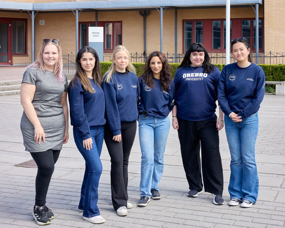 A group of six people wearing casual clothing and navy blue sweatshirts stand together outside a building.