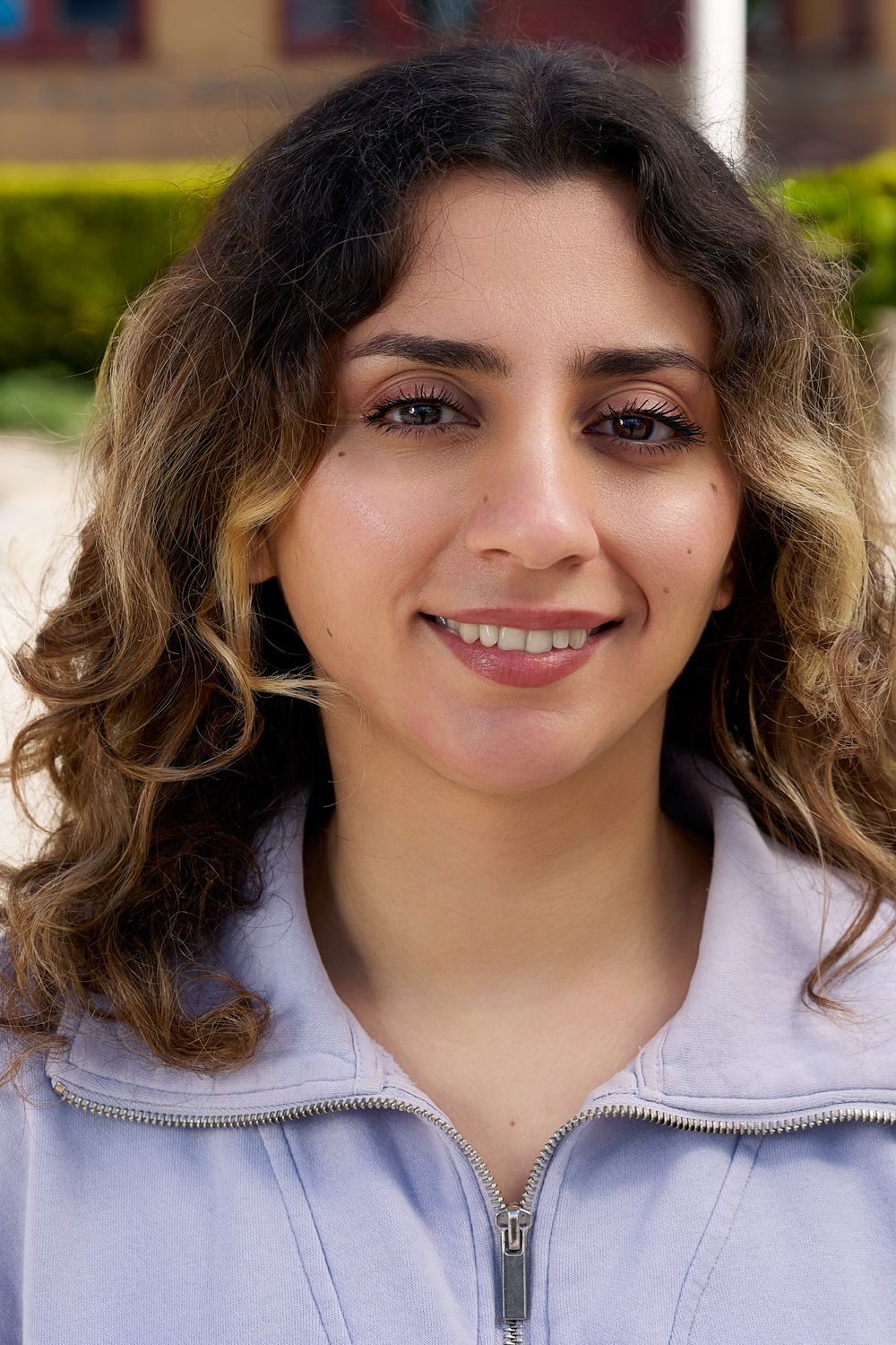 Portrait of someone with curly ombre hair and a friendly smile wearing a light colored zip-up sweater.