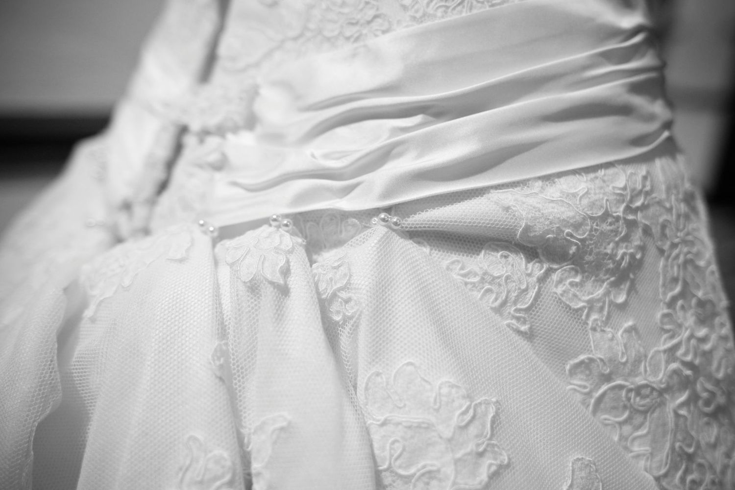 Elegant white wedding dress details showing delicate lace patterns and embroidered fabric in a black and white photograph.