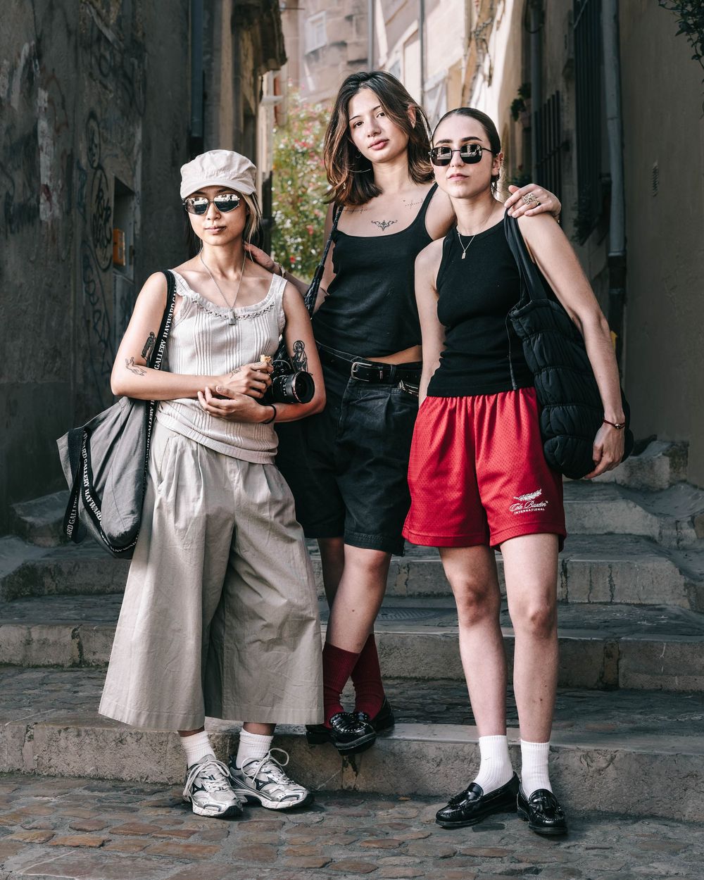 Three stylish friends in urban streetwear pose together on historic stone steps in a narrow alley.