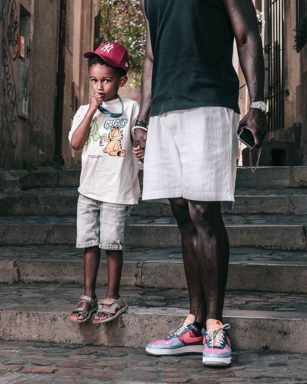 Child wearing a red cap and casual outfit stands on stone steps next to an adult in a dark shirt and white shorts.