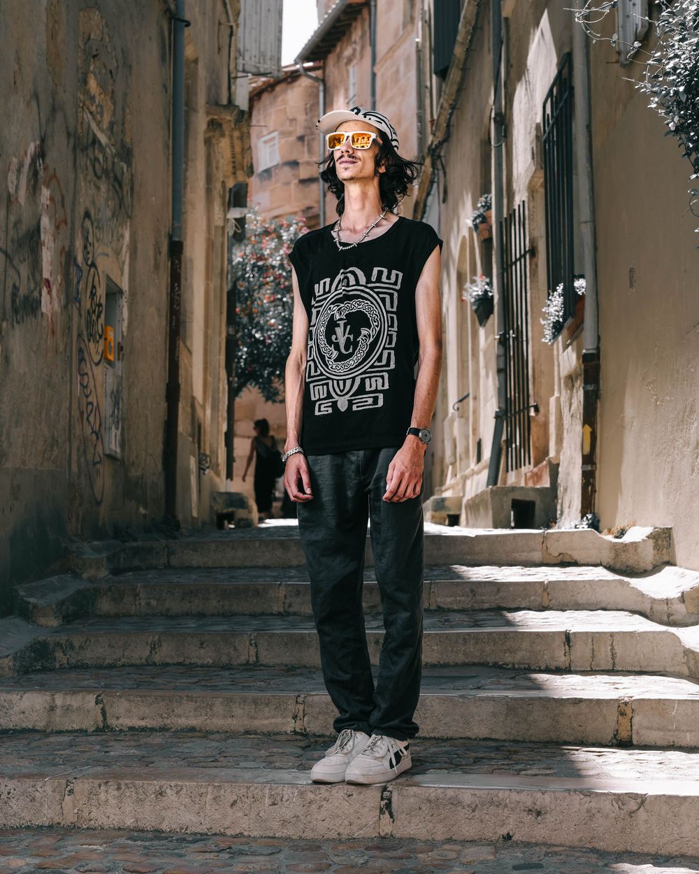 Figure in a rock band t-shirt and jeans stands on stone steps in an old European alleyway in sunlight.