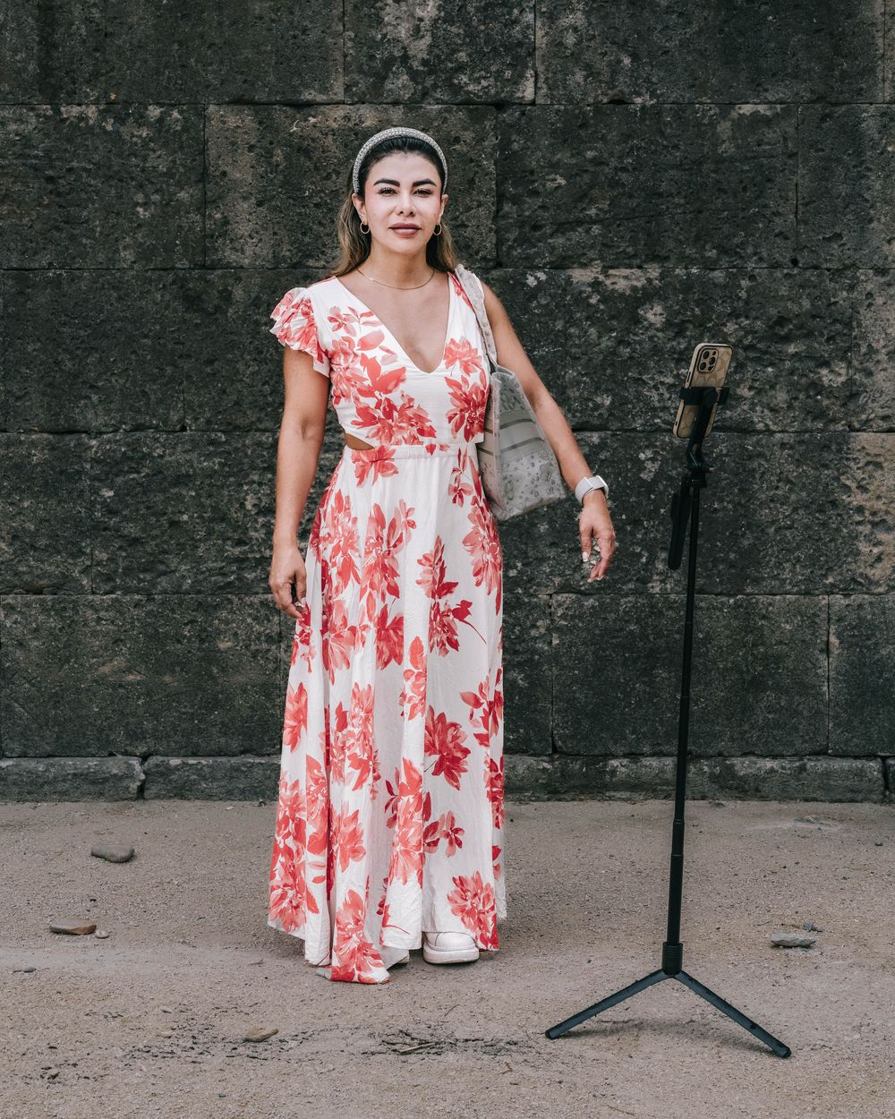A woman in a flowing red and white floral maxi dress stands against a textured stone wall near a microphone stand.