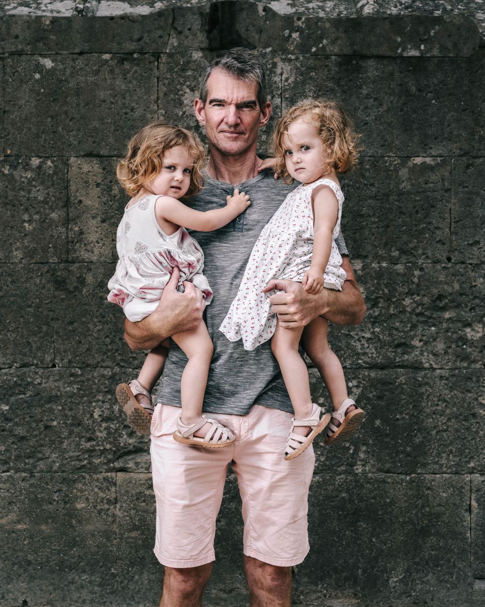 A person holds two young children wearing light colored summer outfits against a dark stone wall background.