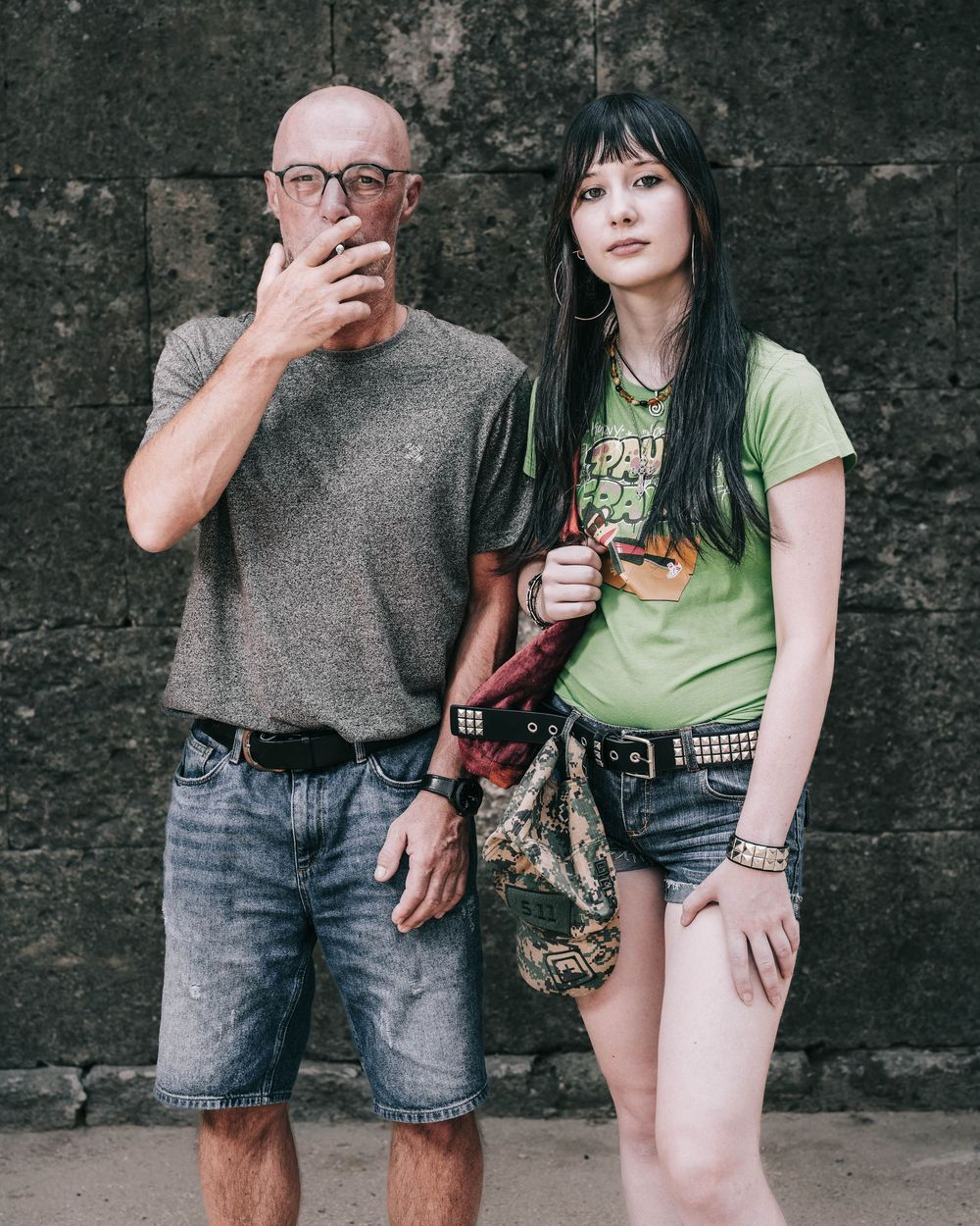 Two people posing against a dark wall, one wearing a grey top and shorts, the other in a green graphic t-shirt.