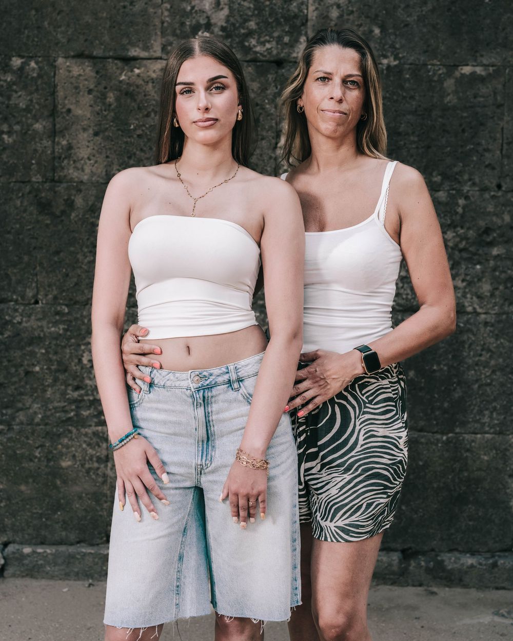 Two people in white tops and light denim standing against a textured dark wall.