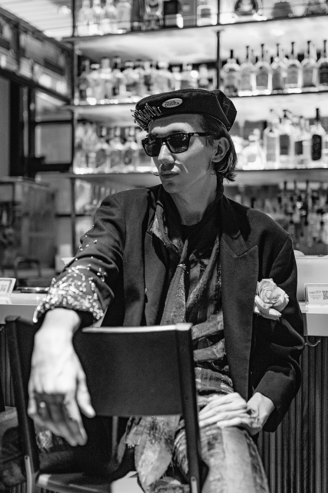 A black and white photo of someone in sunglasses and a cap sitting at a bar with liquor bottles in the background.