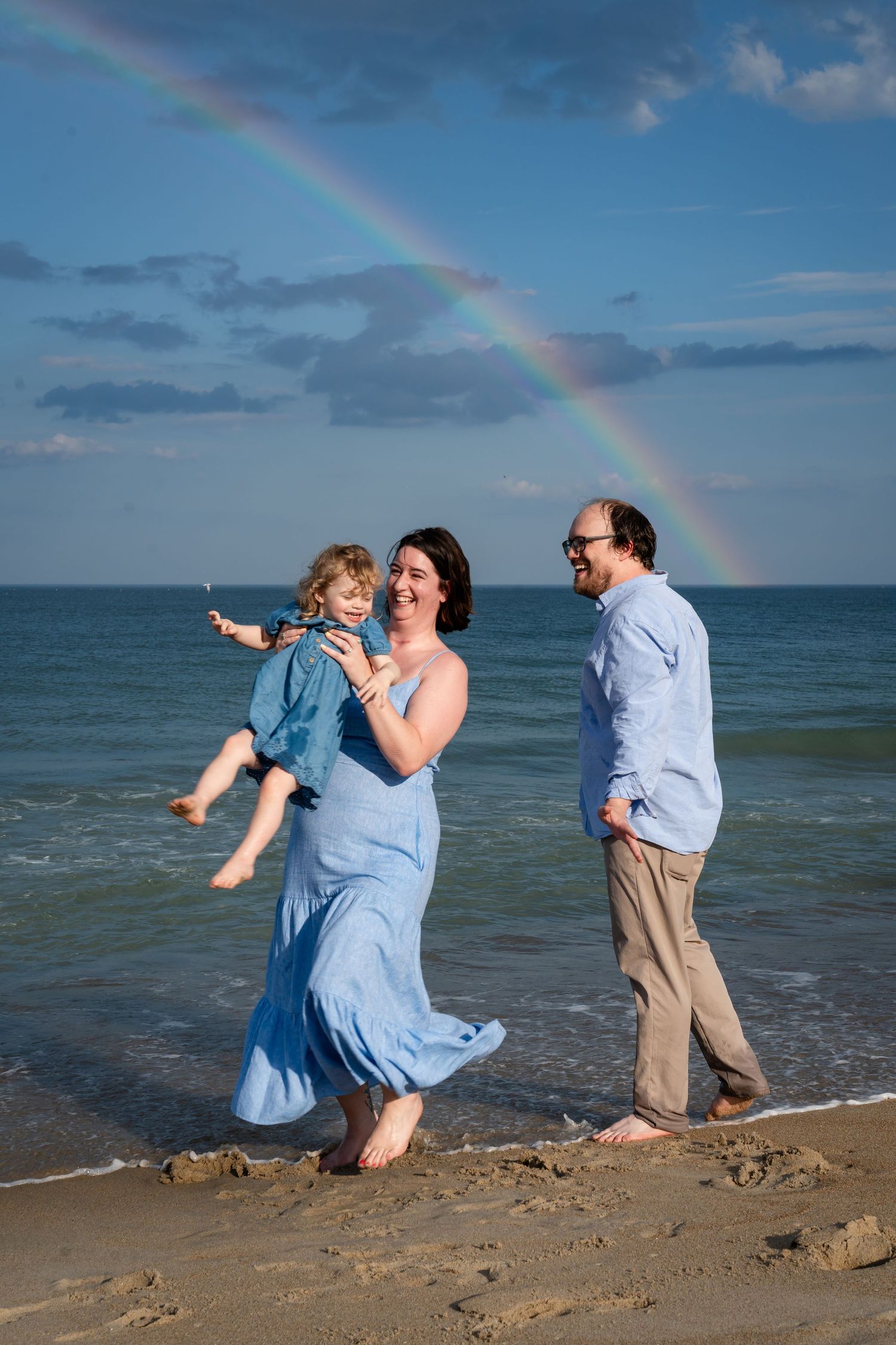 A family enjoying a playful moment on the beach with a vibrant rainbow arching across the blue sky over the ocean.