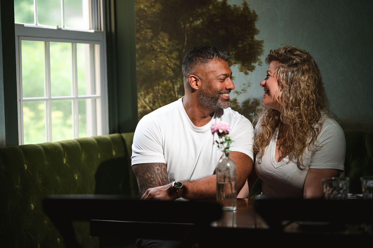 A couple shares an intimate moment while sitting together on a green velvet couch by a window.