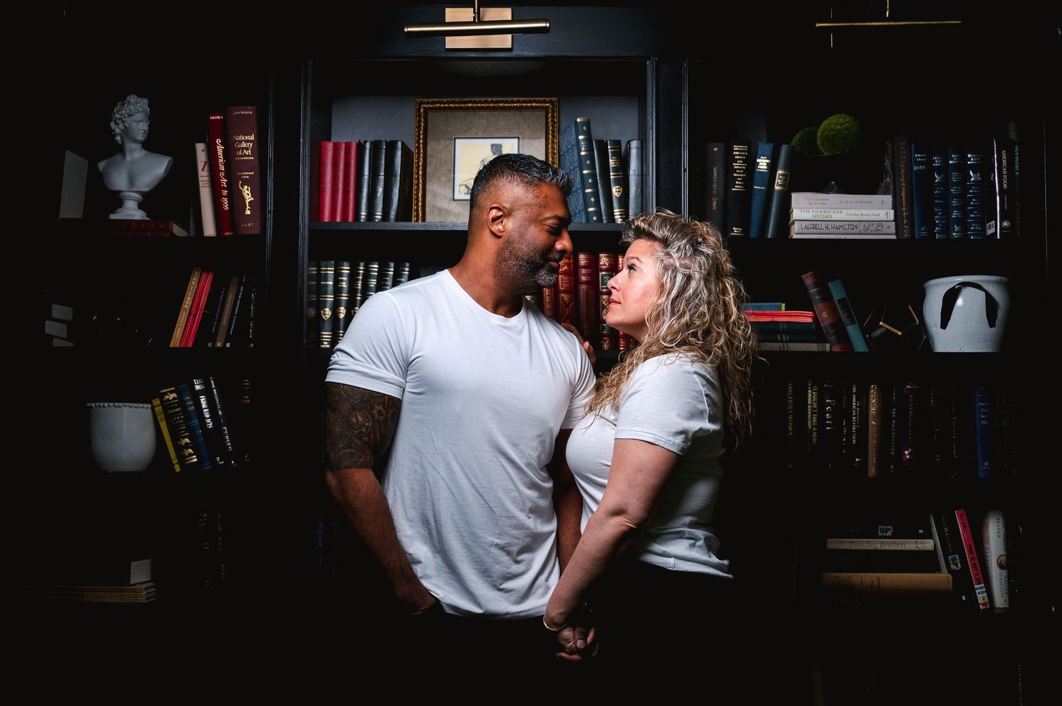 A romantic couple embraces in a moody library setting with bookshelves behind them.