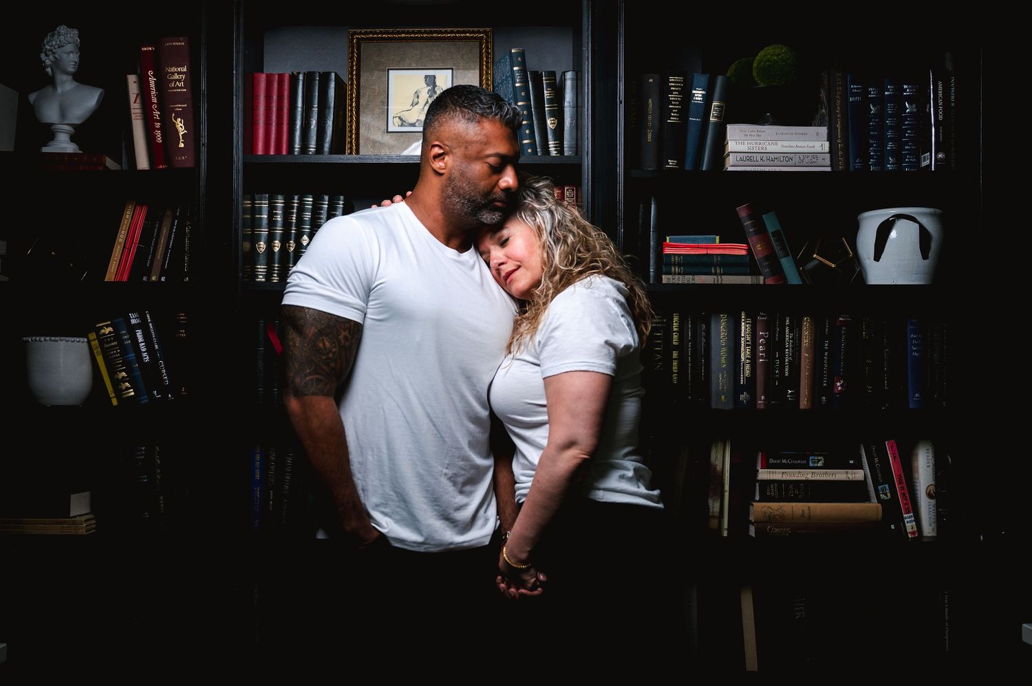 A tender moment between two people sharing an embrace in front of a dark bookshelf filled with books and decor.