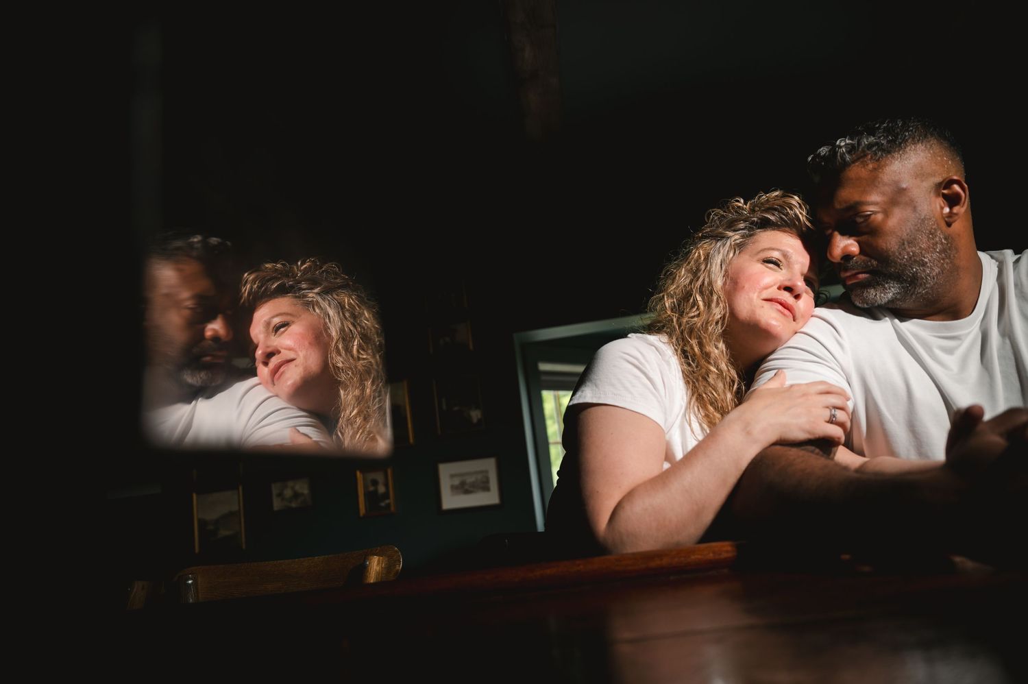 Two people share an intimate moment while sitting at a dark wooden table in a dimly lit space.