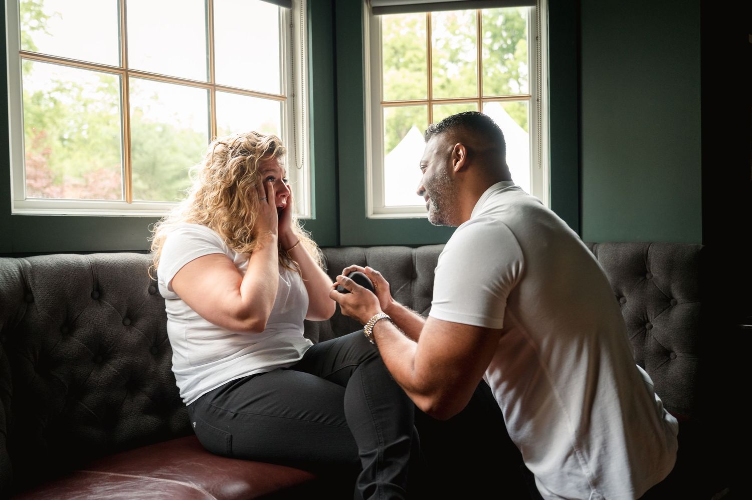 Two people sitting on a dark gray tufted couch by a window share an emotional moment in a room with green walls.