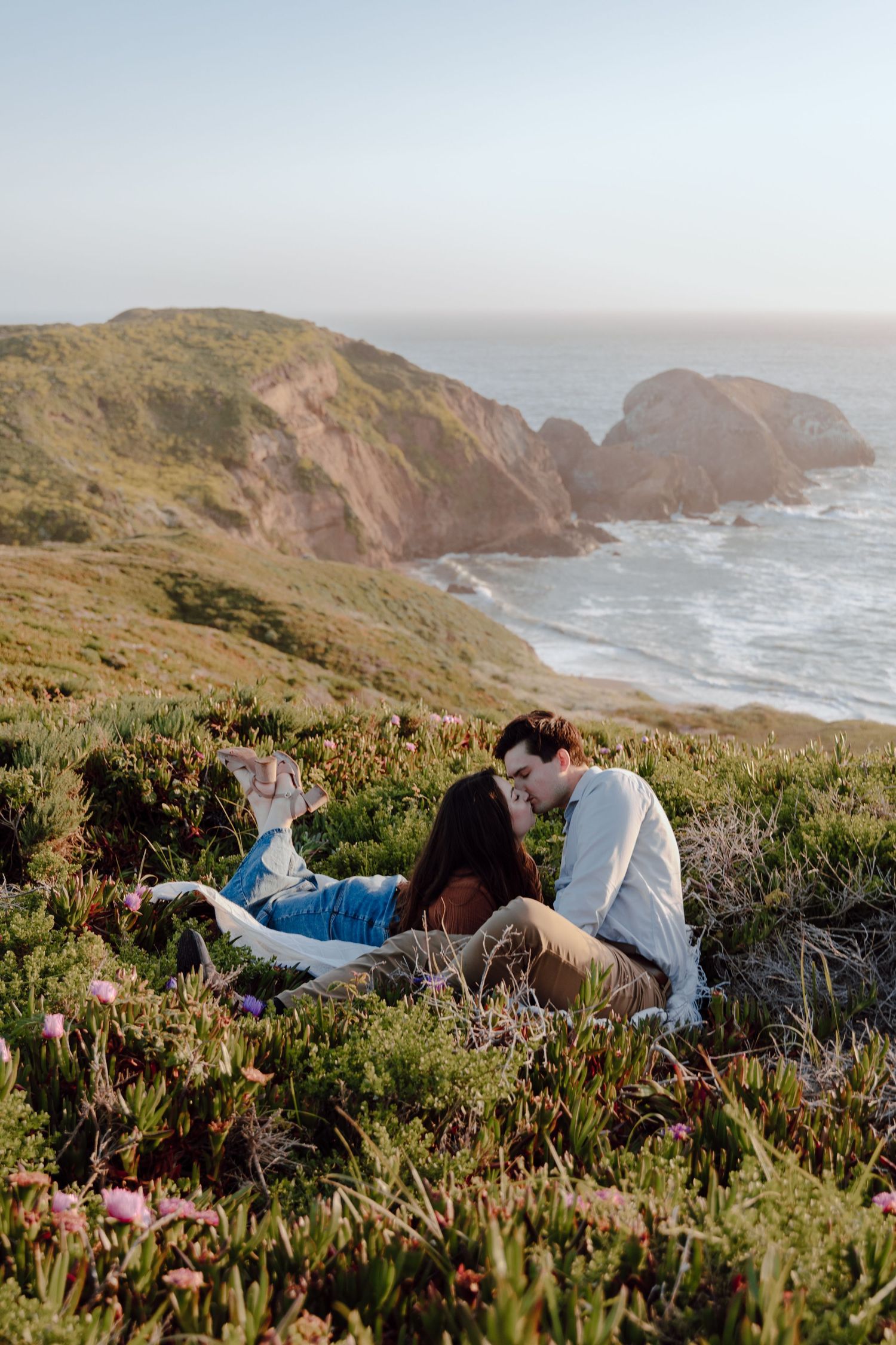 A couple enjoys a scenic picnic overlooking dramatic coastal cliffs and ocean waves during golden hour.