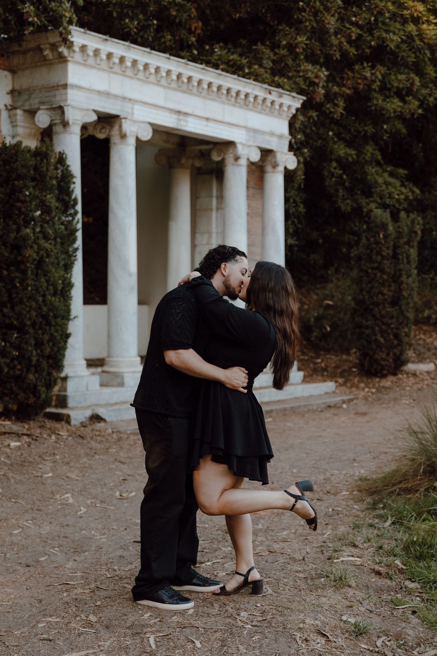 A romantic couple embraces near a white classical columned garden structure in a warm outdoor setting.