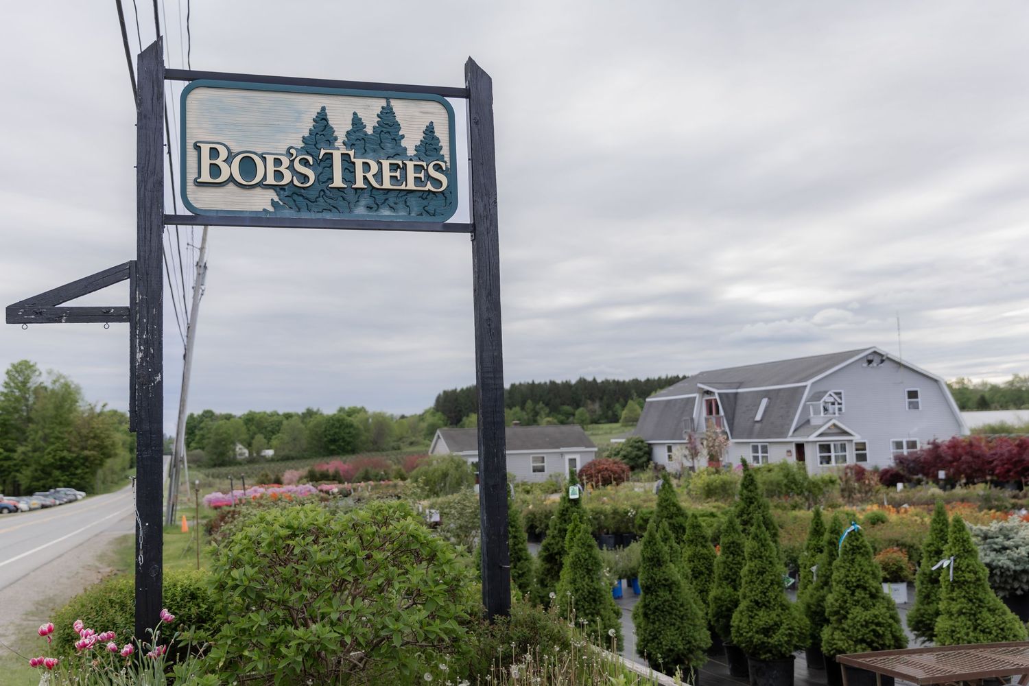 A sign for Bob's Trees stands in front of a garden center and nursery on a cloudy day.
