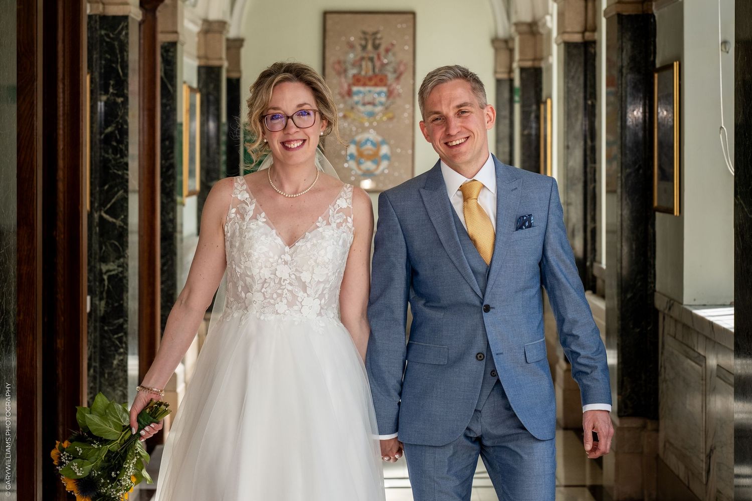 A happy couple walking together at their wedding, with the bride in white lace and groom in blue suit with yellow tie.