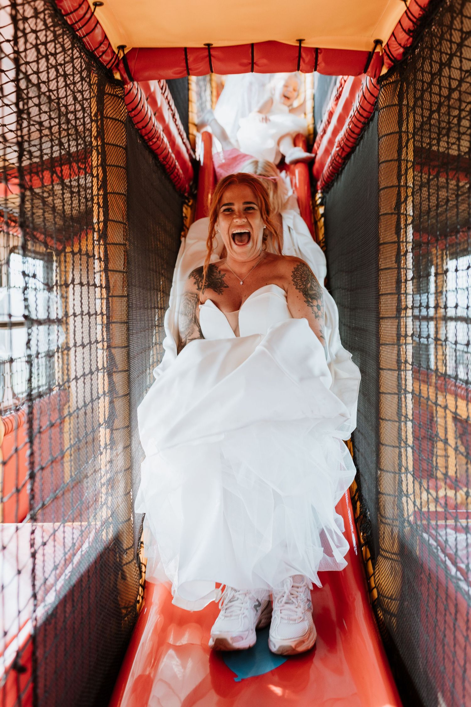 Bride wearing a flowy white wedding dress slides down an indoor playground slide with joyful excitement.