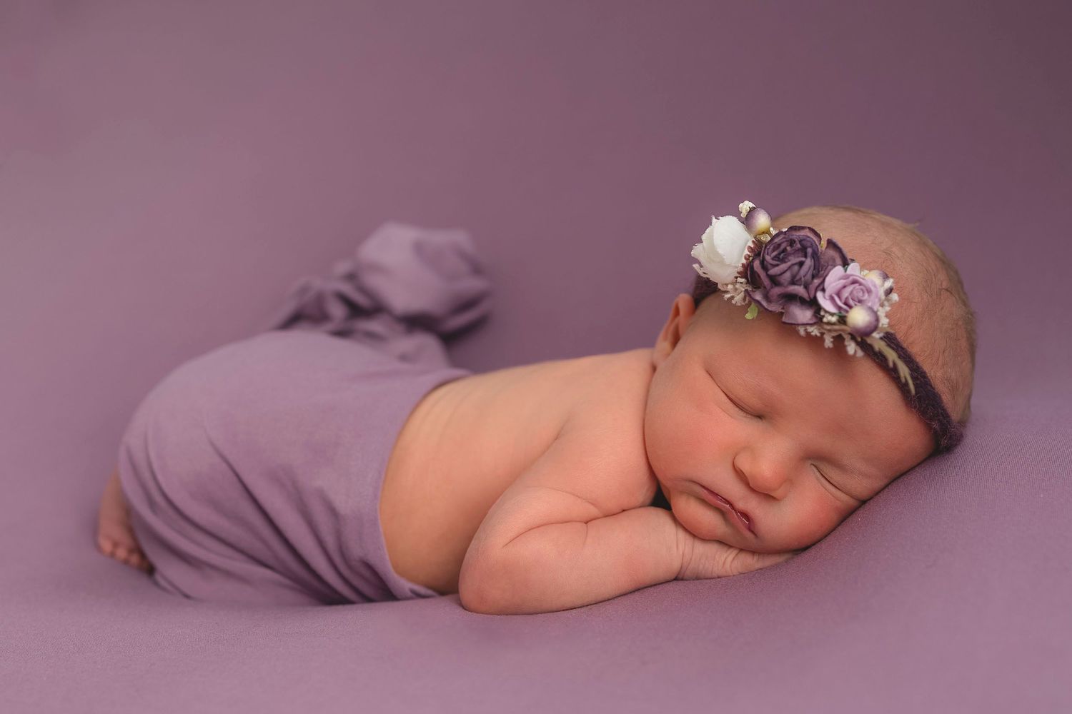 A sleeping newborn wearing purple floral headband poses on purple backdrop during photography session. Hampshire newborn baby photography