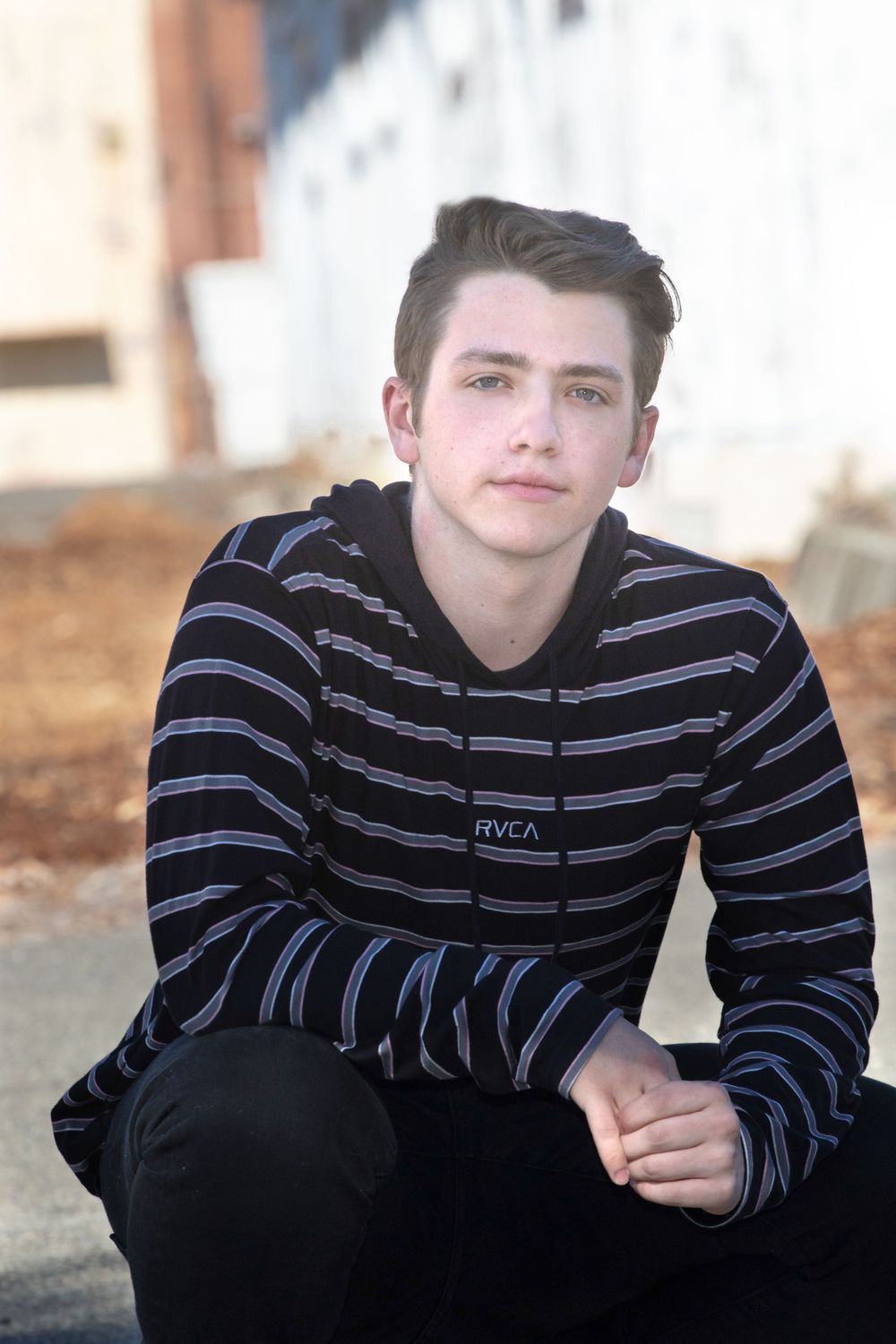 Young person in striped black hoodie and dark pants poses casually outdoors against blurred background. Senior portrait photo