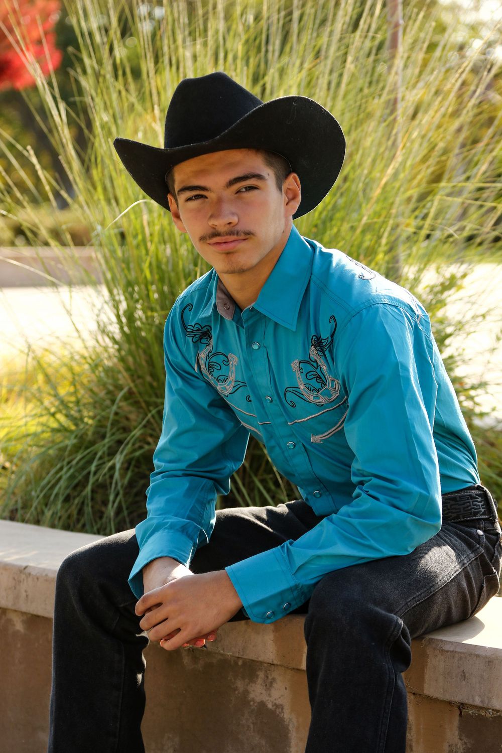 A person in Western attire with a black cowboy hat and turquoise embroidered shirt sits casually on a concrete ledge. Senior portrait photo
