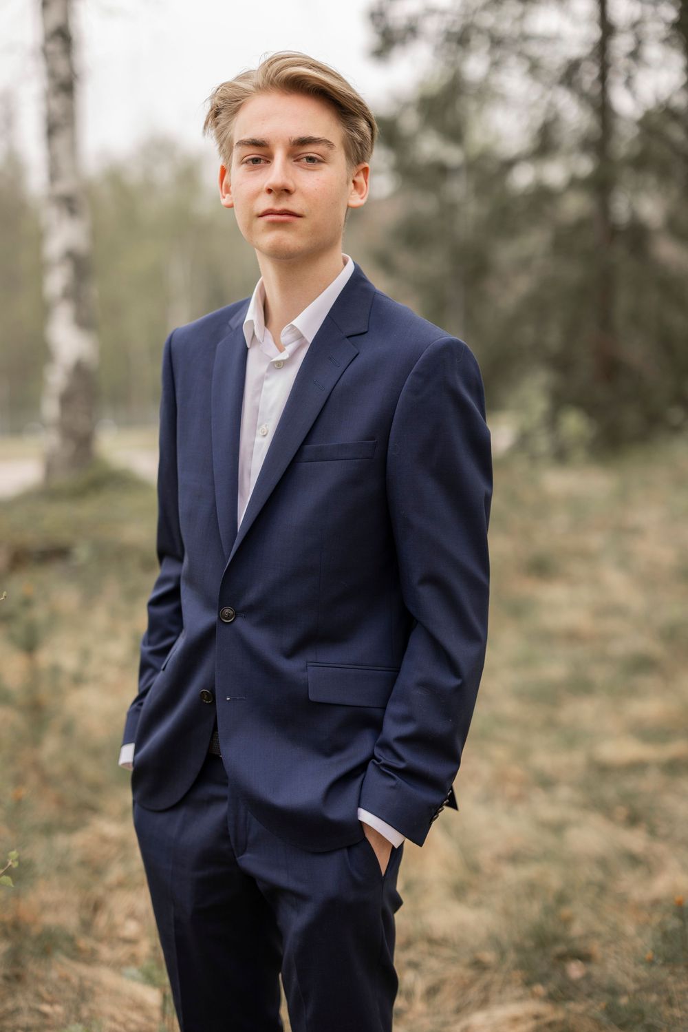 A well-dressed person in a navy blue suit and white shirt poses outdoors in a wooded setting on a cloudy day.