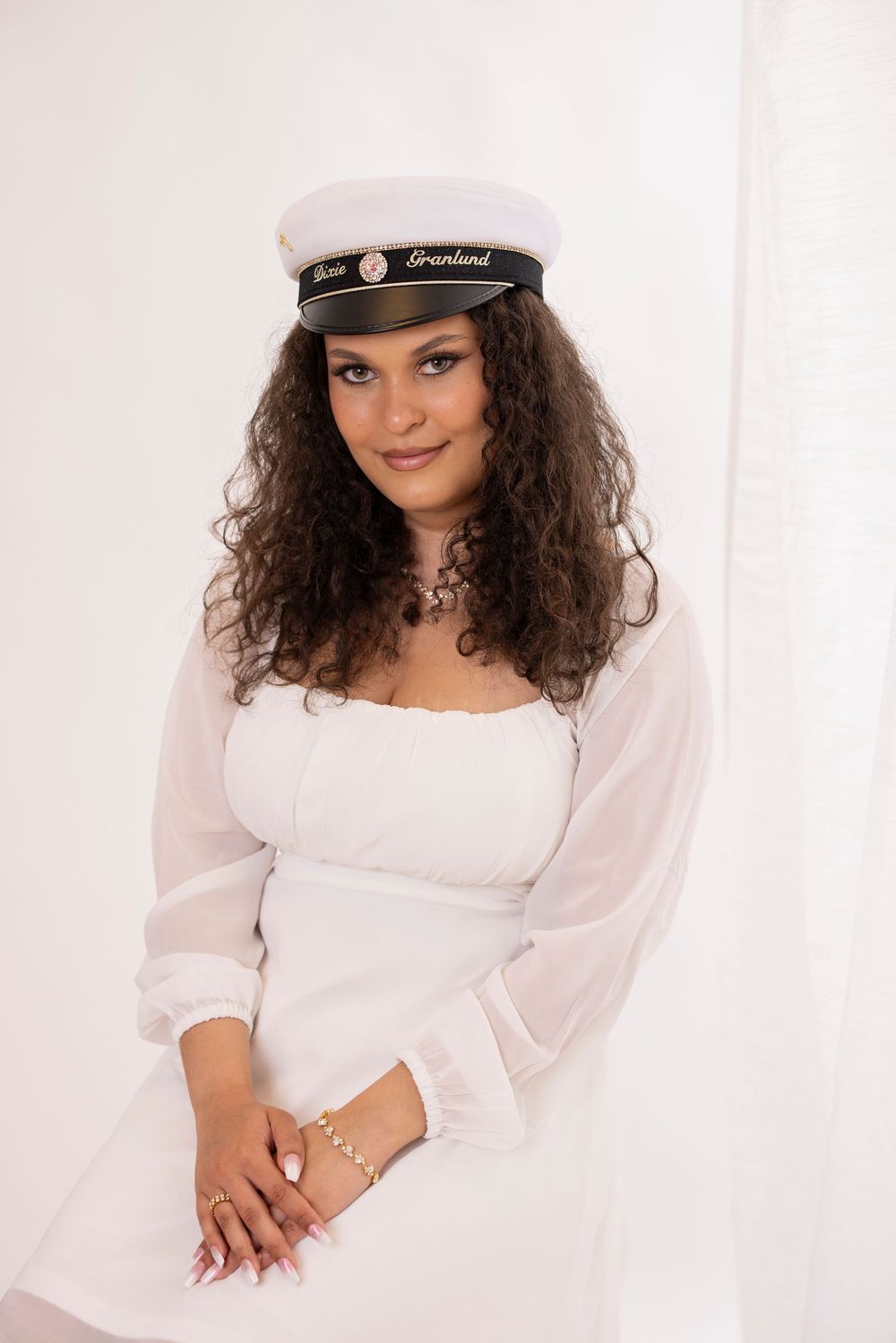 A model in a white flowing dress and sailor's cap smiles gracefully against a light backdrop.