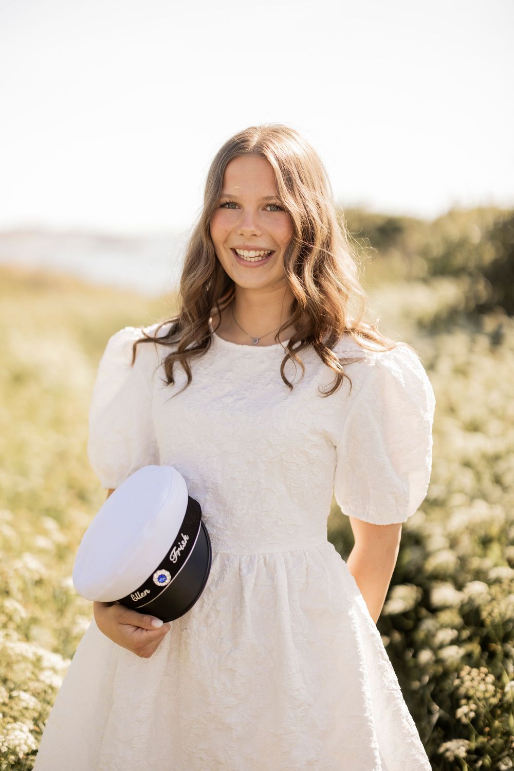 A woman in a white puff-sleeve dress stands in a sunny meadow holding a motorcycle helmet.
