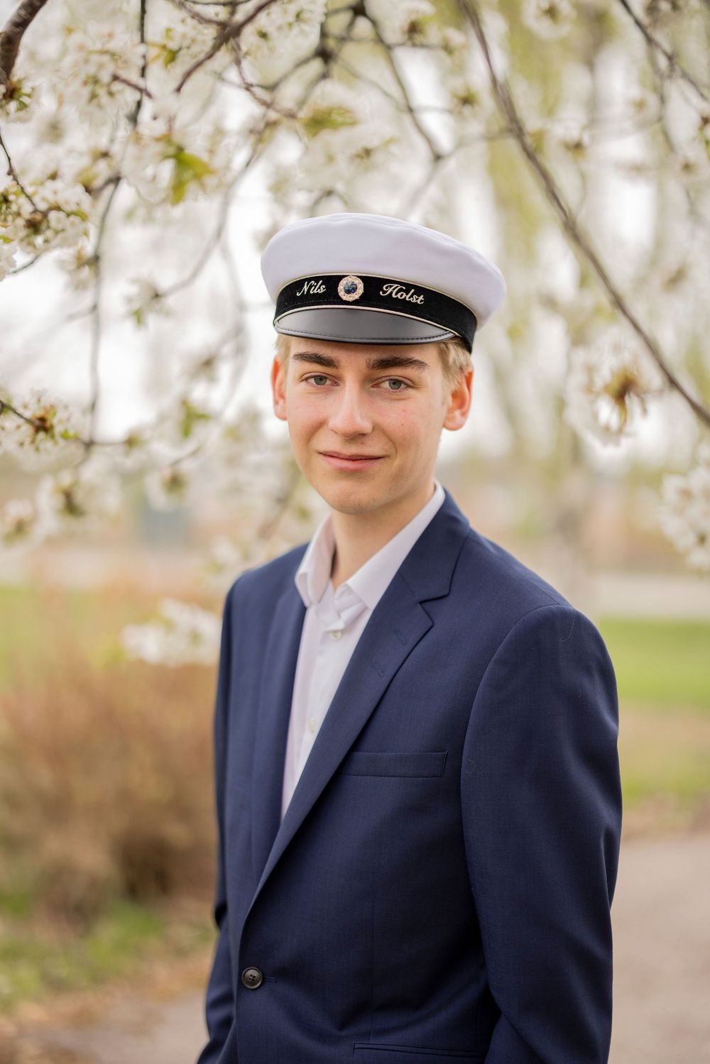 A student in navy blazer and white sailor cap poses outdoors in front of blooming spring trees.