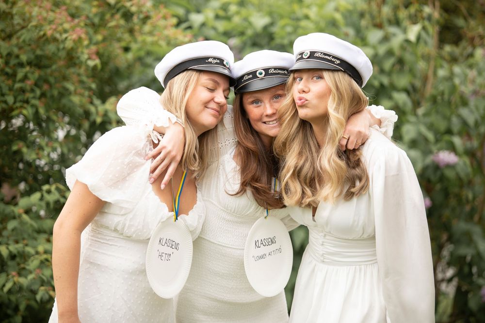 Three companions in white dresses and sailor caps share a joyful moment together in a garden setting.