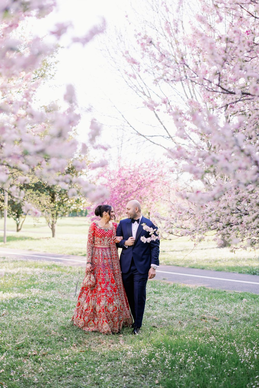 A couple strolls together under blooming cherry blossom trees, with the person wearing a traditional red embroidered gown.