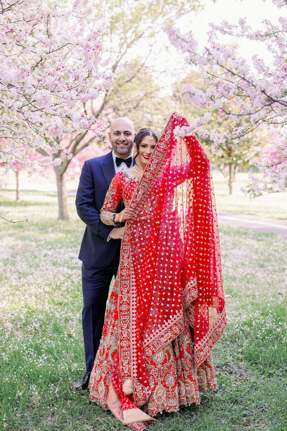 A couple poses under blooming cherry blossom trees, with traditional wedding attire including a red embroidered sari.