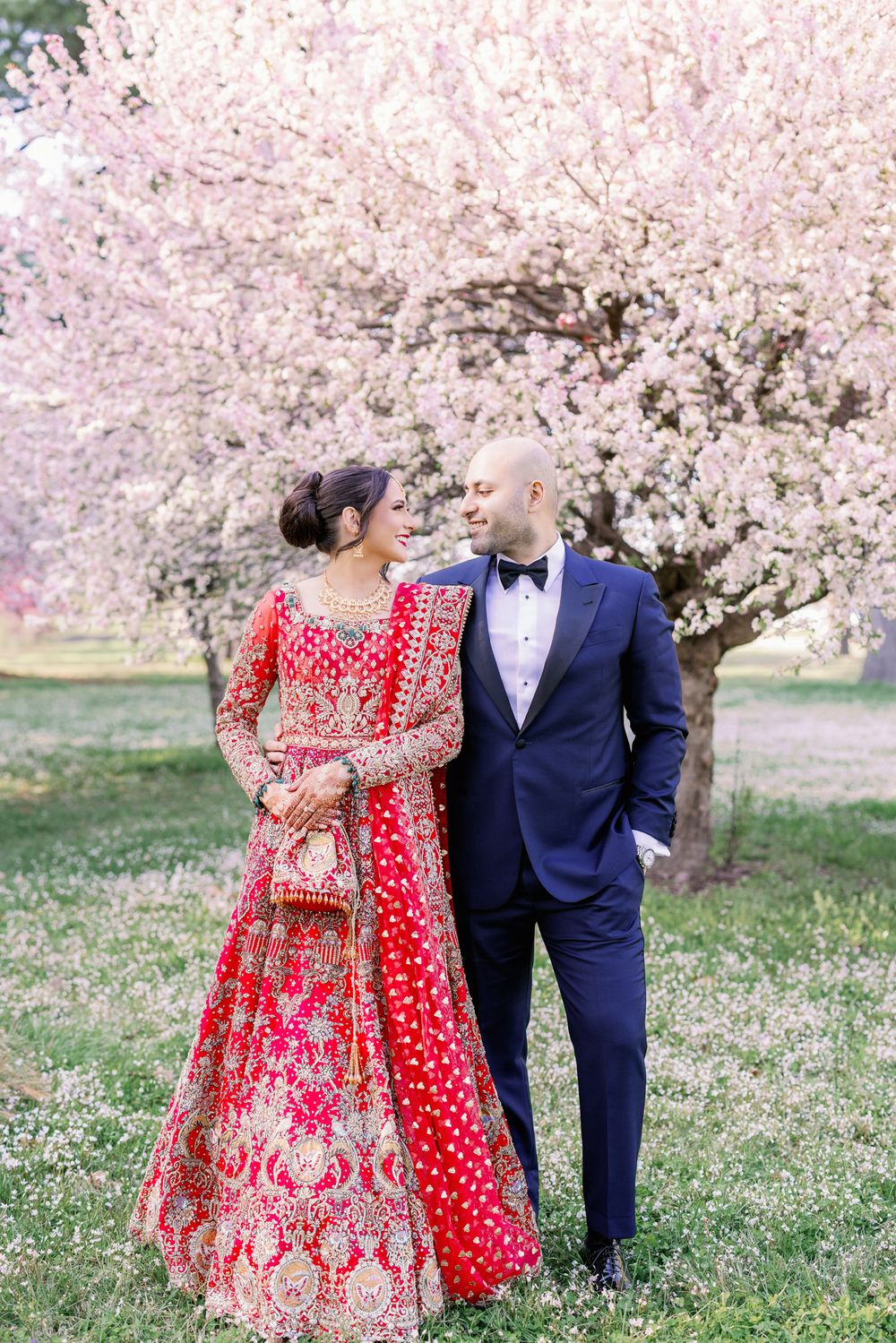 A couple in traditional wedding attire stands beneath blooming cherry blossom trees in spring.