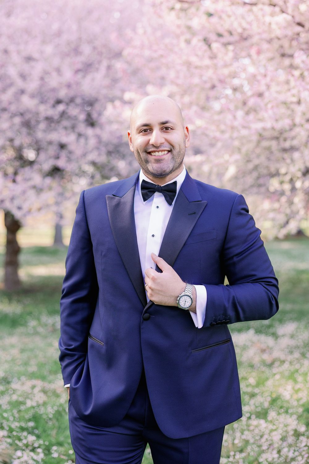 A person in a navy blue tuxedo with bow tie poses outdoors during cherry blossom season.
