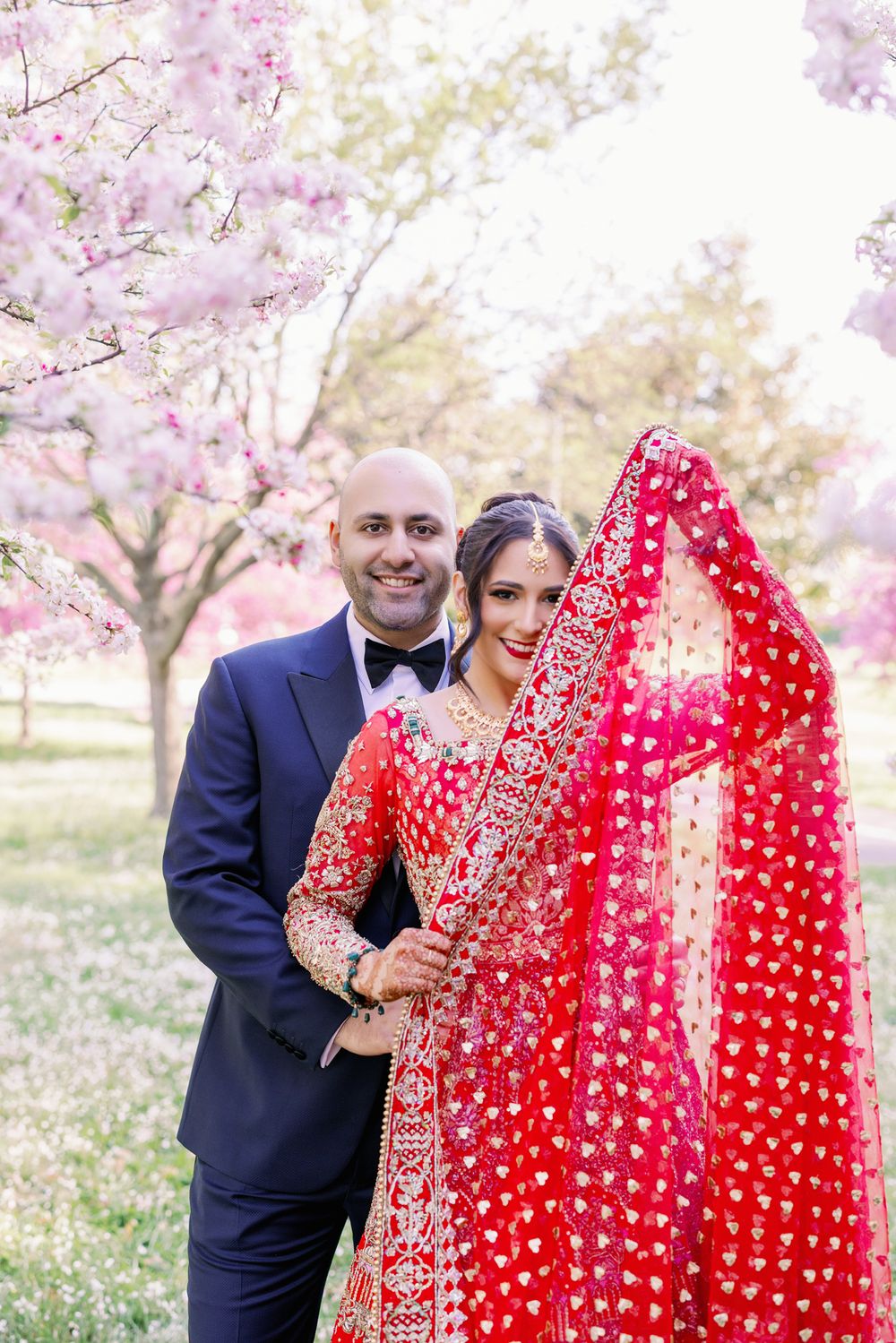 A couple in traditional wedding attire stands beneath blooming cherry blossom trees in a spring garden setting.