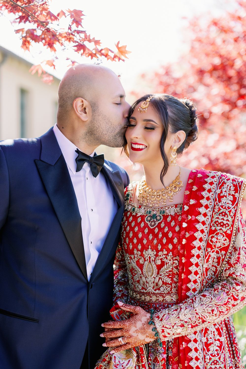 A romantic couple in traditional wedding attire posing under vibrant red autumn maple leaves.