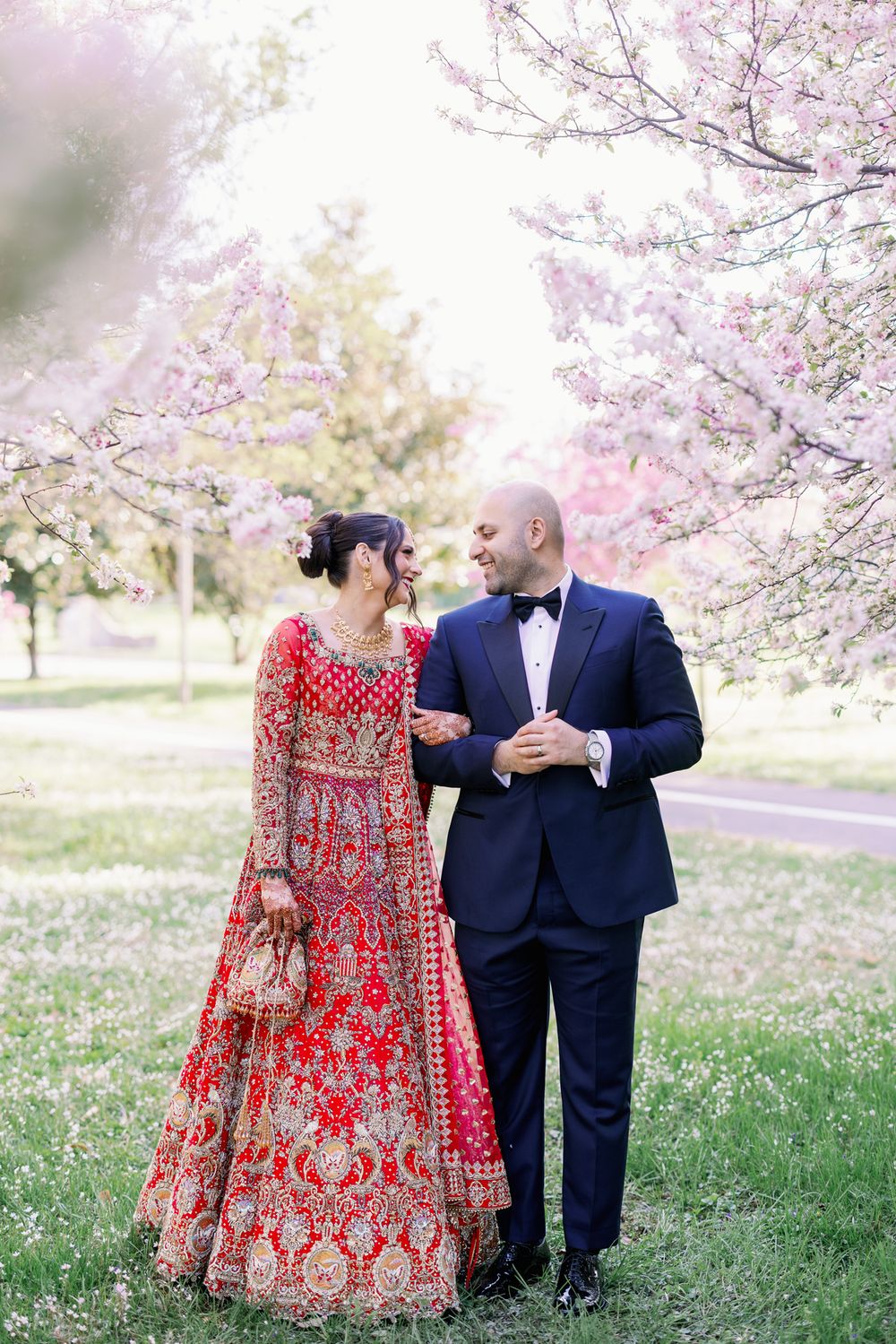 A couple in wedding attire standing together under blooming cherry blossom trees in a spring garden.