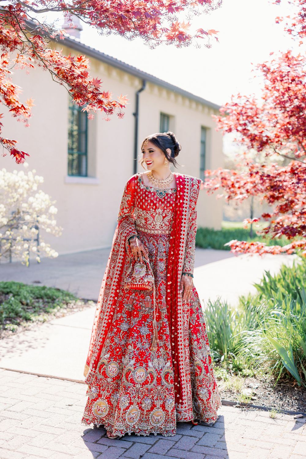 A traditional red and gold embroidered lehenga worn outdoors amid blooming pink cherry blossom trees.