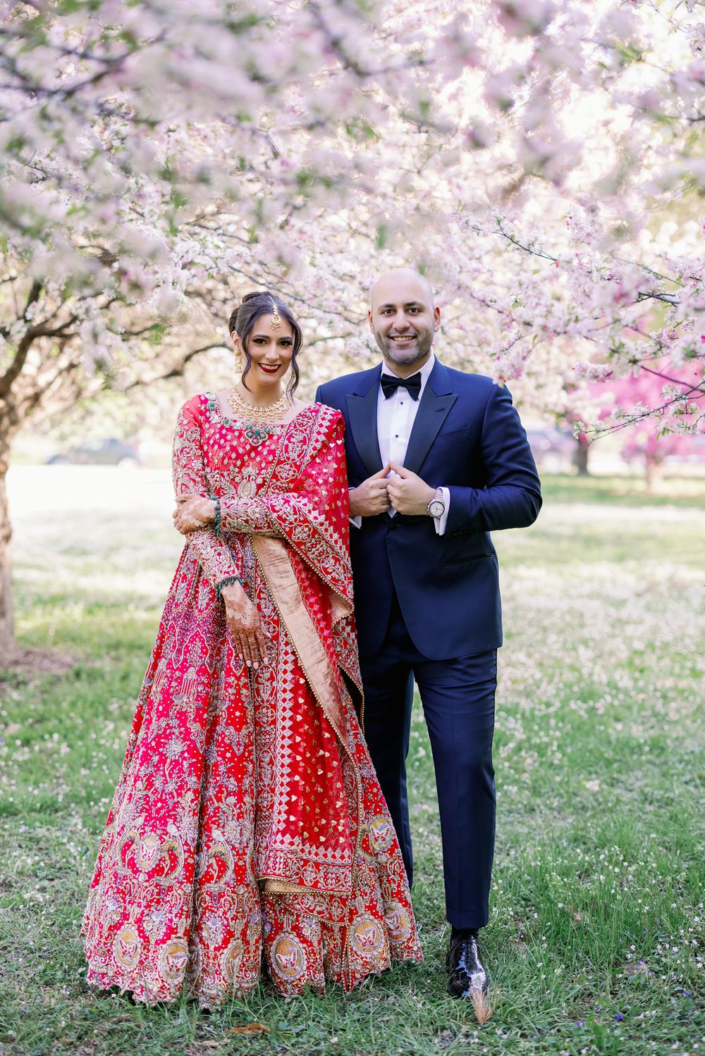 A couple poses under blooming cherry blossoms, with one person in a traditional red lehenga and another in a navy suit.