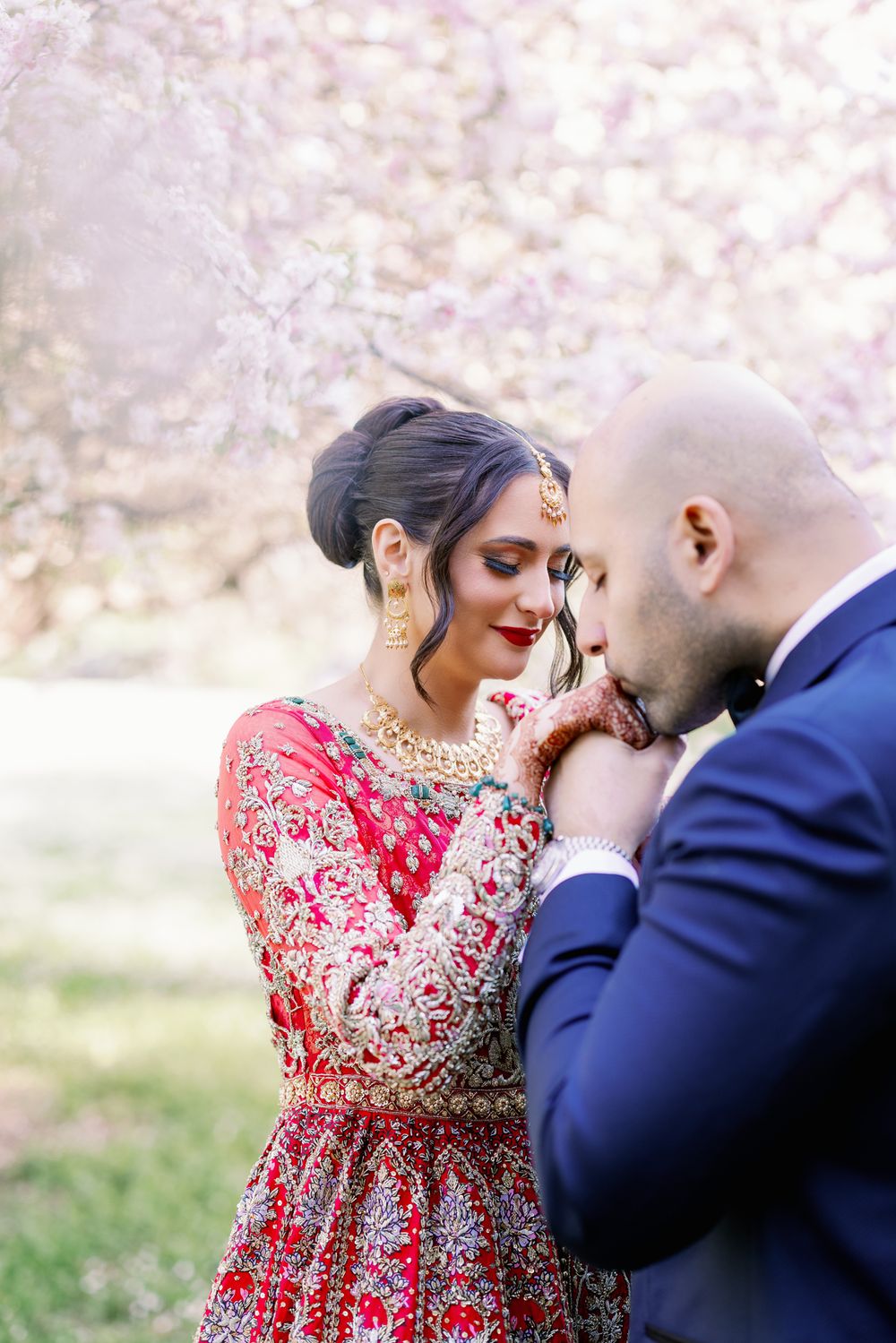 A romantic couple shares an intimate moment outdoors, with the bride wearing an ornate red and gold embroidered gown.