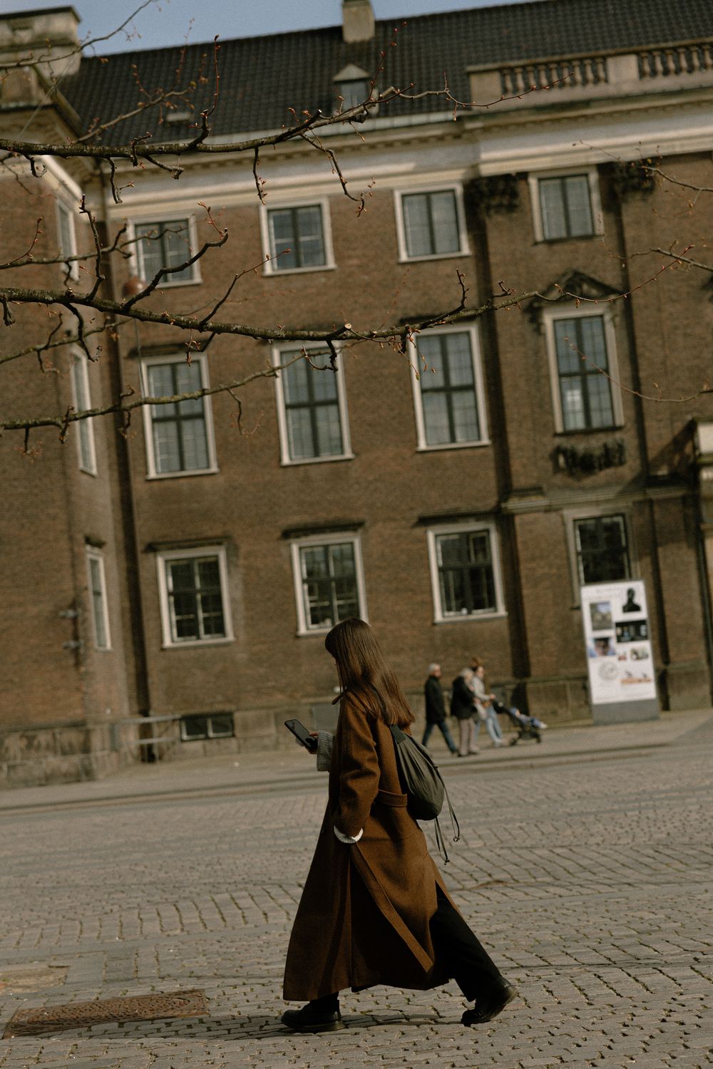Person in long brown coat walking past historic building facade on cobblestone street. Destination wedding photographer Copenhagen