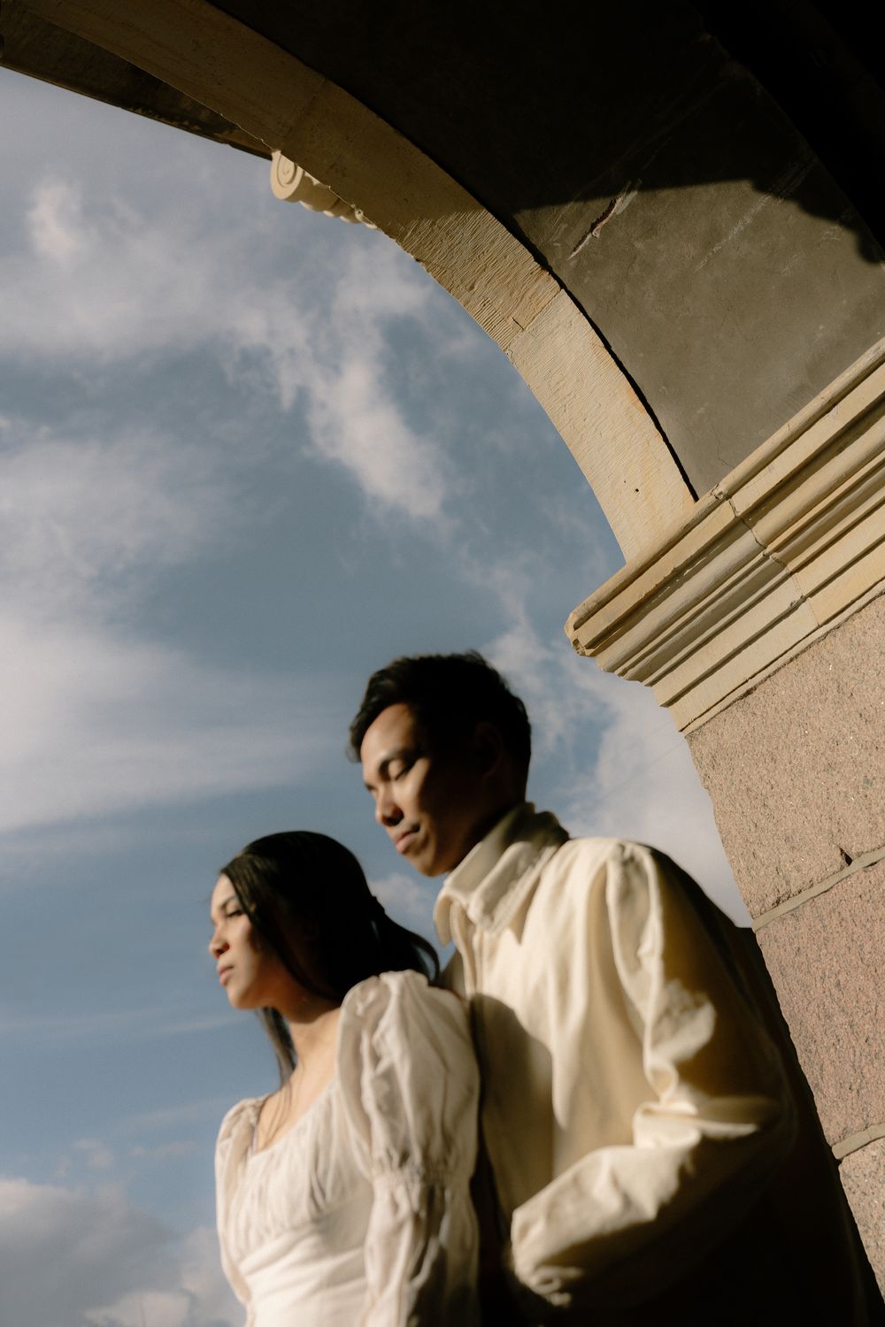 Romantic couple in white outfits pose under an architectural arch against cloudy sky. Destination wedding photographer Copenhagen