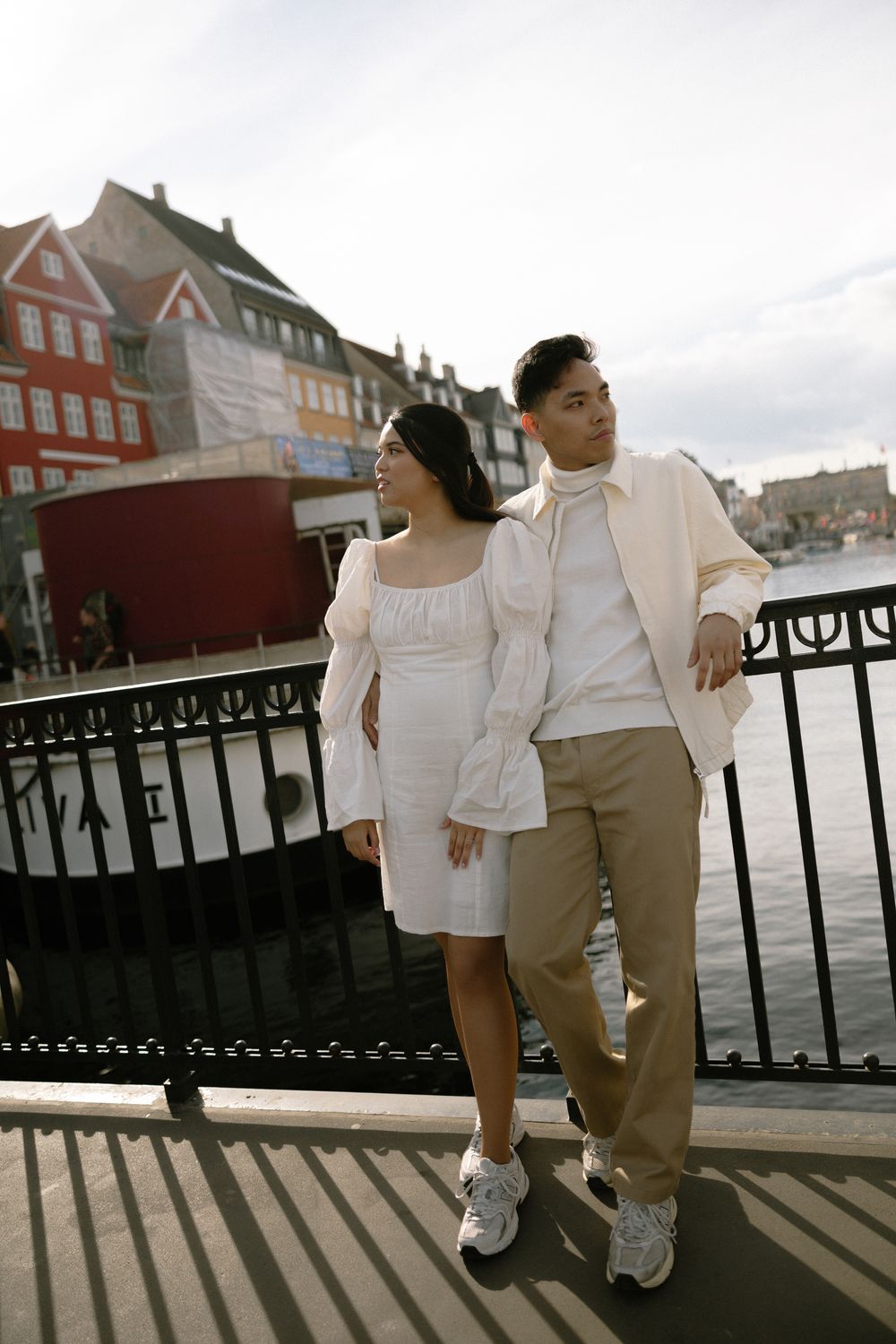 Stylish couple in white outfits pose together on waterfront with colorful buildings behind. Destination wedding photographer Copenhagen