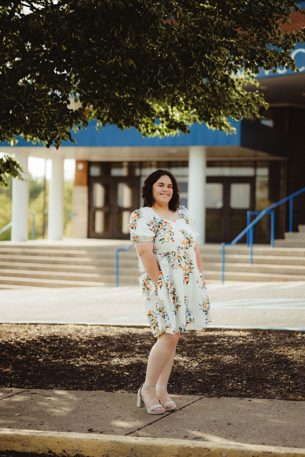 A person in a white floral dress and heels stands on a sidewalk near building steps under a leafy tree.