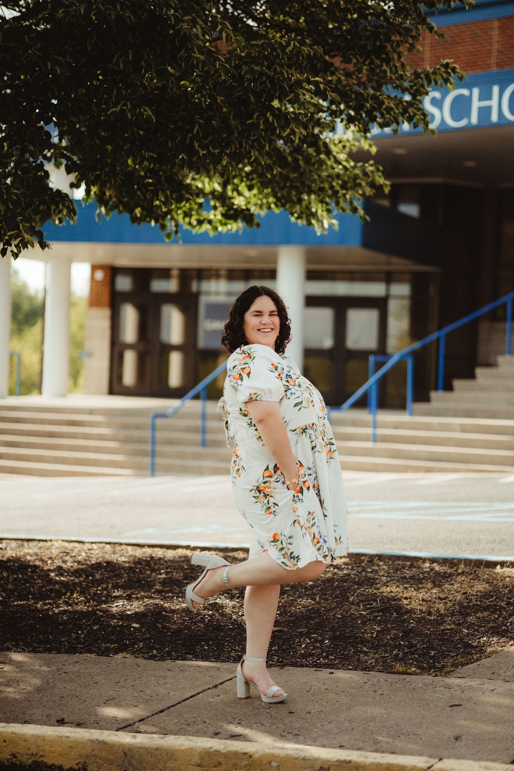A person in a floral dress and heels poses in front of a school building on a sunny day.