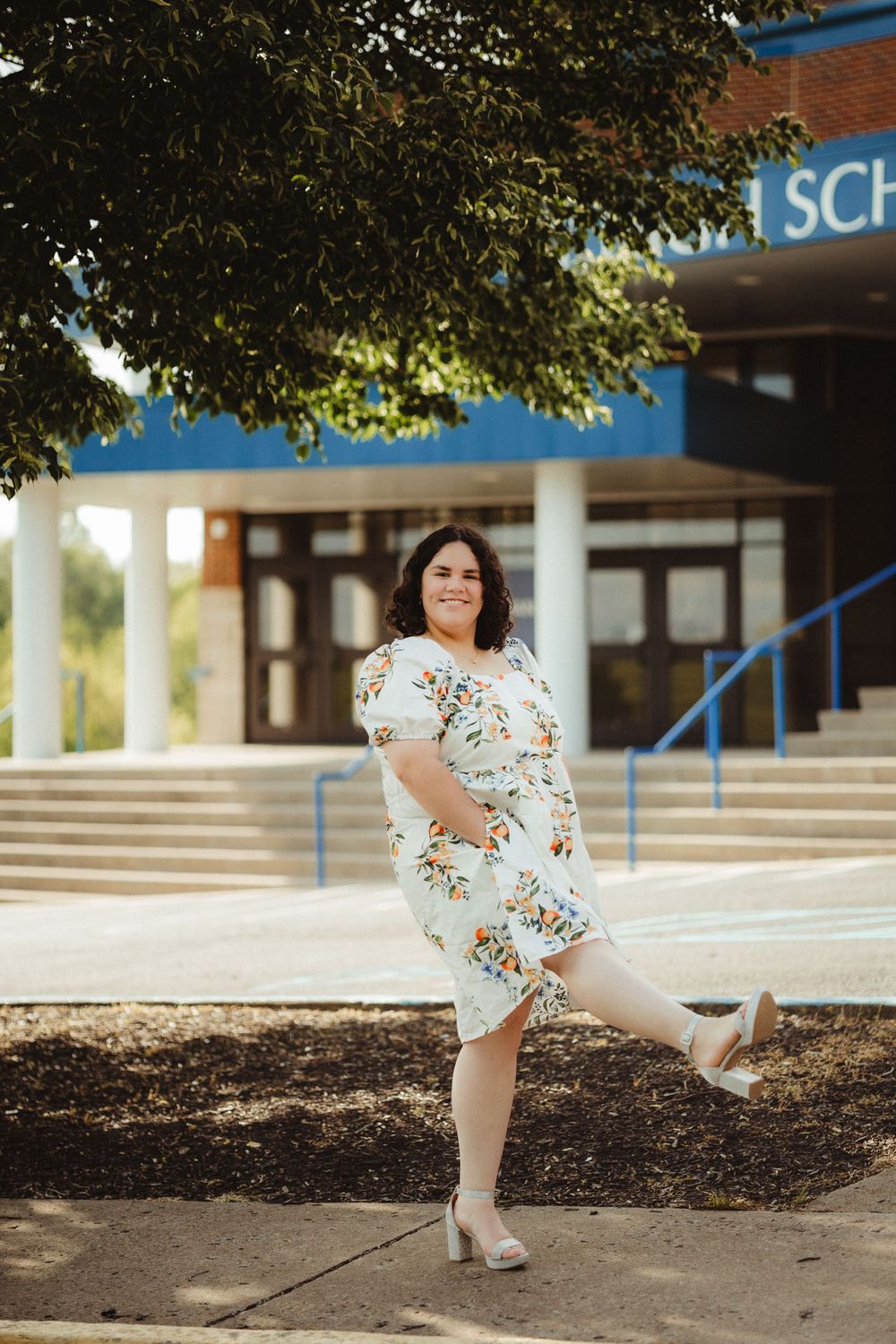 A person in a floral white dress poses playfully outside a school building on a sunny day.