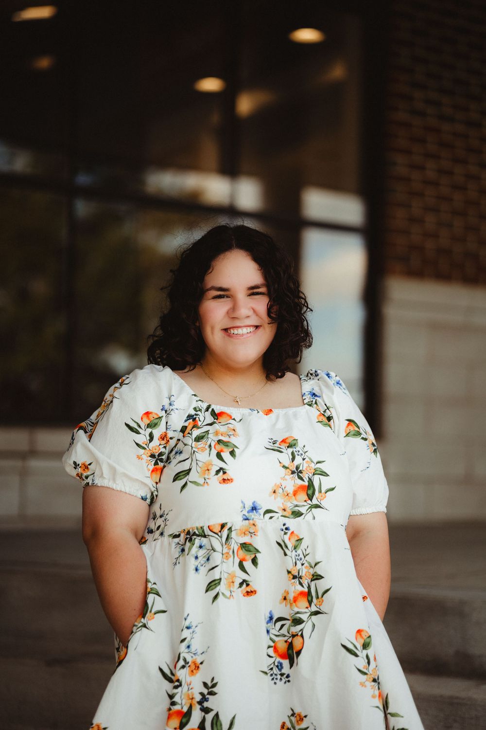 A person wearing a white floral dress with orange flowers poses outside a brick building in warm lighting.
