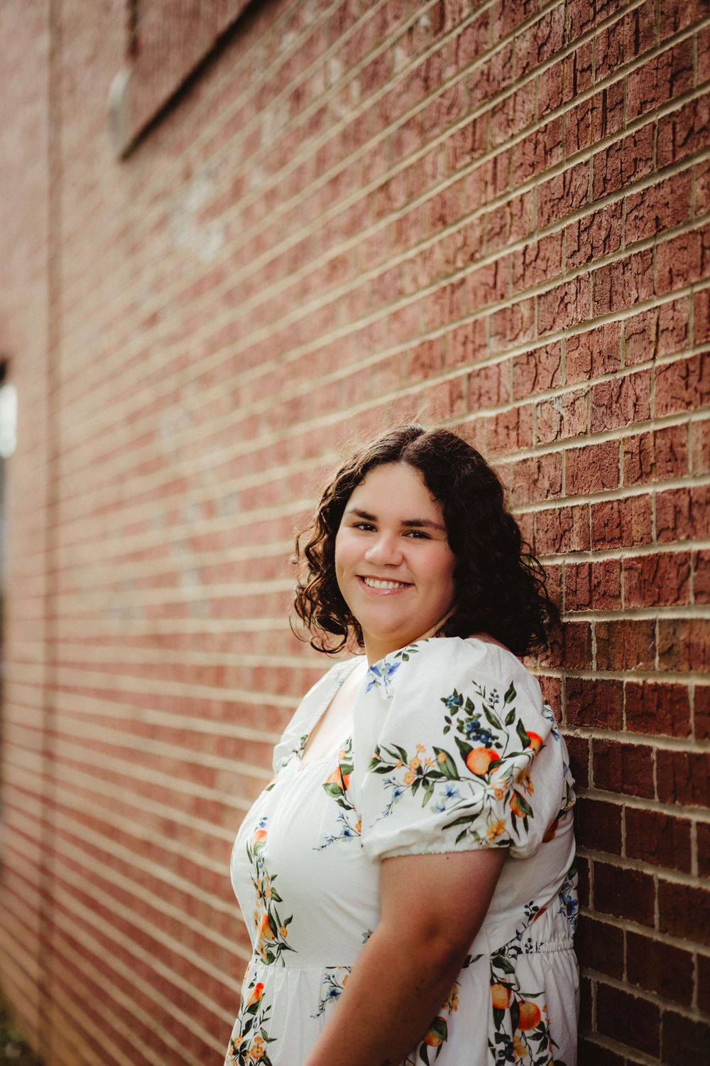 A person in a white floral dress stands against a rustic brick wall while smiling at the camera.