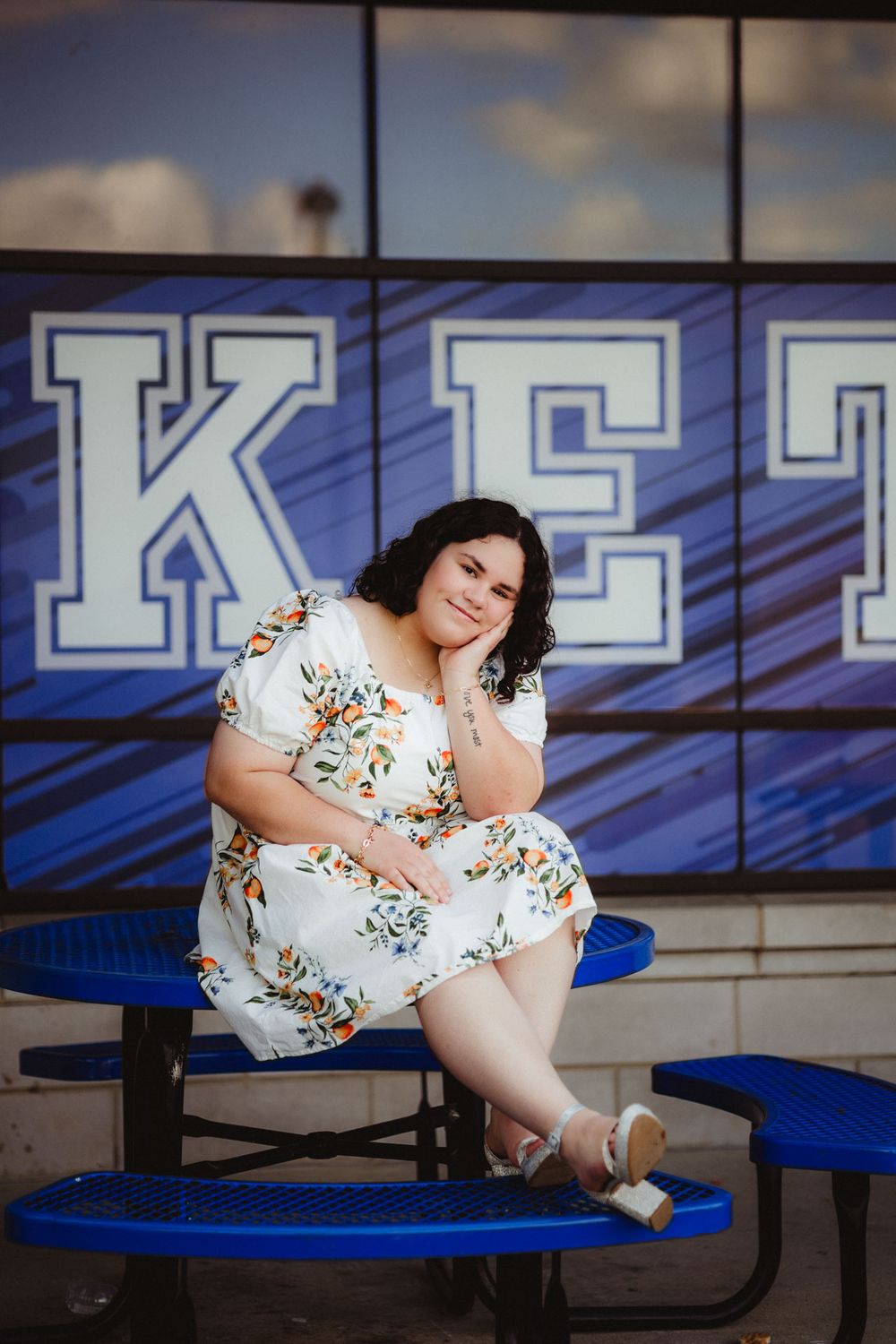 A person in a white floral dress poses on blue bleachers in front of large block letters reading KE.