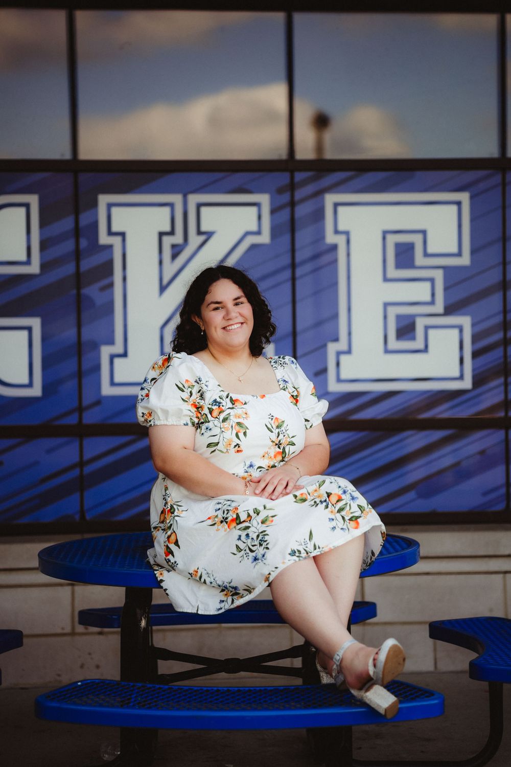 A person in a floral white dress sits on blue bleachers on a sunny day with large letters visible in the background.
